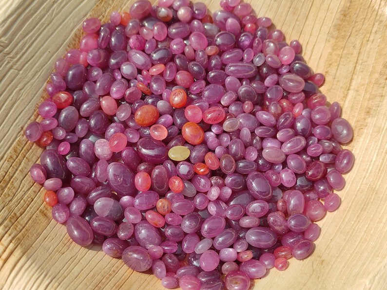 May include: A collection of polished, oval-shaped gemstones in shades of purple, pink, and red. The gemstones vary in size and are scattered on a light brown, textured surface. The gems have a smooth, glossy appearance.