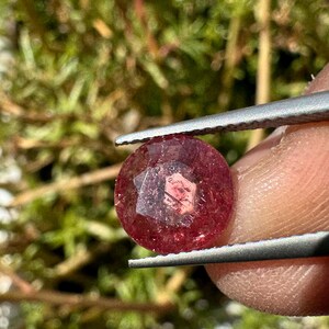 May include: A round, faceted, red gemstone with a clear center and a pink hue. The gemstone is held in tweezers.