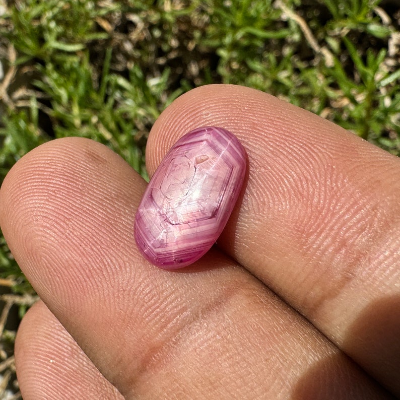 May include: A pink star ruby gemstone with a hexagonal facet pattern. The gemstone is oval shaped and has a smooth, polished surface.
