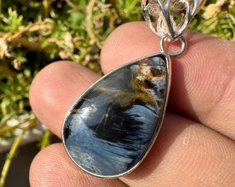 Puede incluir: Un colgante de ojo de tigre azul en forma de lágrima con detalles plateados. El colgante está engastado en una argolla plateada y tiene una cadena plateada.