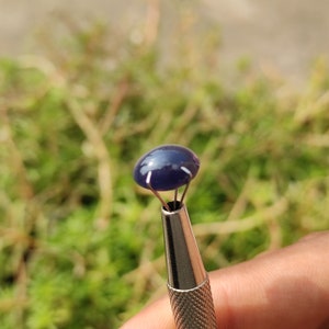 May include: A close-up of a dark blue oval gemstone held in a metal tool. The gemstone is smooth and shiny, with a slight sparkle.