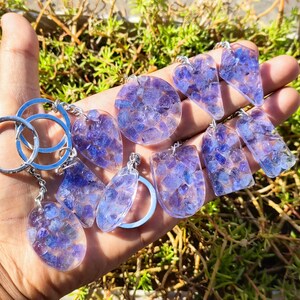 May include: A set of 10 purple and clear resin keychains with a variety of shapes, including hearts, squares, and circles. Each keychain features a unique design of small purple crystals embedded in the resin.