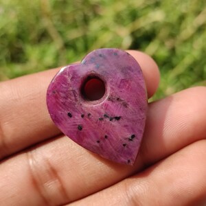 May include: A heart-shaped pendant made of pink ruby with black inclusions. The pendant has a hole drilled through the top for stringing.