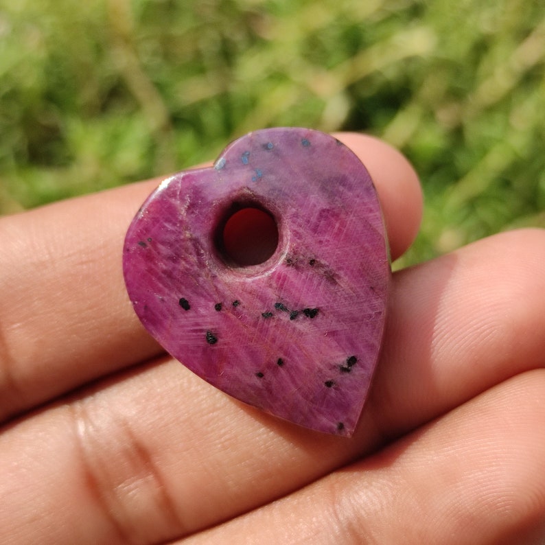 May include: A heart-shaped pendant made of pink ruby with black inclusions. The pendant has a hole drilled through the top for stringing.