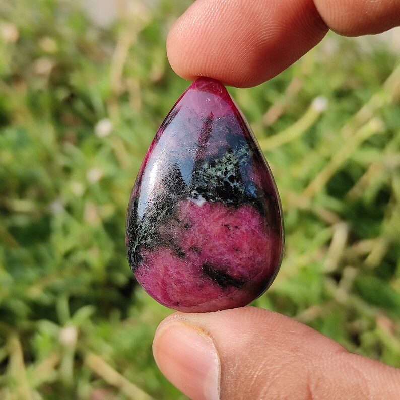 Puede incluir: Un colgante de piedra cabuj&oacute;n en forma de l&aacute;grima con un patr&oacute;n rosa y negro. La piedra tiene un acabado brillante y est&aacute; engastada en un engaste de tono plateado.