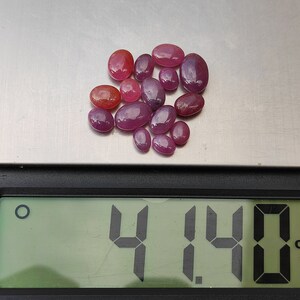 May include: A collection of oval-shaped, polished gemstones in shades of red and pink are displayed on a digital scale. The scale reads 4.14 ct, indicating the total carat weight of the gemstones. The gemstones vary in size and color intensity.
