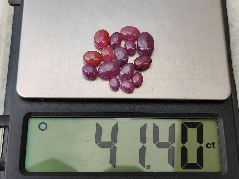 May include: A collection of oval-shaped, polished gemstones in shades of red and pink are displayed on a digital scale. The scale reads 4.14 ct, indicating the total carat weight of the gemstones. The gemstones vary in size and color intensity.