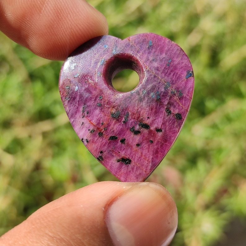 May include: A heart-shaped pendant made of pink ruby with black inclusions. The pendant has a hole in the center for a chain or cord.
