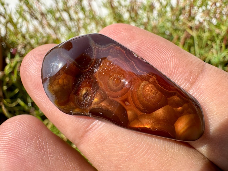 May include: A polished fire agate gemstone with a smooth, irregular teardrop shape. The stone features deep translucent brown and amber tones with iridescent, glowing orb-like inclusions that shimmer in shades of orange and gold under light.