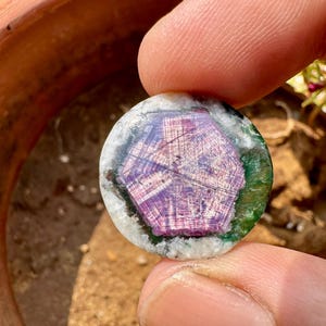 K&ouml;nnte beinhalten: Ein runder Edelstein mit einem zentralen sechseckigen Muster in Lila- und Rosat&ouml;nen, umgeben von Gr&uuml;n und Wei&szlig;. Der Edelstein wird zwischen zwei Fingern gehalten und zeigt seine polierte Oberfl&auml;che und nat&uuml;rlichen Farben.