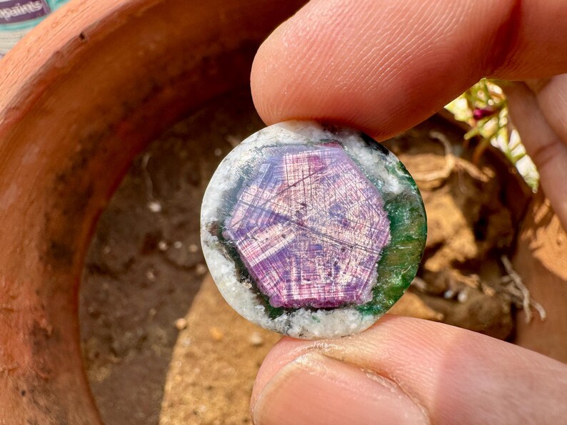 K&ouml;nnte beinhalten: Ein runder Edelstein mit einem zentralen sechseckigen Muster in Lila- und Rosat&ouml;nen, umgeben von Gr&uuml;n und Wei&szlig;. Der Edelstein wird zwischen zwei Fingern gehalten und zeigt seine polierte Oberfl&auml;che und nat&uuml;rlichen Farben.