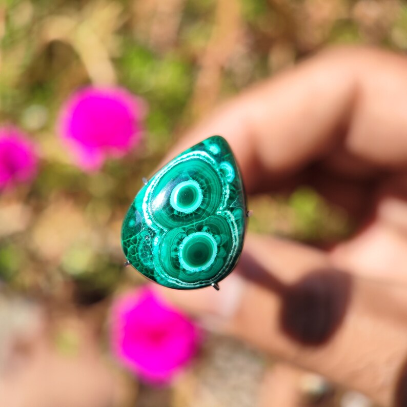 May include: A teardrop-shaped green malachite cabochon with a swirling pattern of concentric circles. The stone is set in a silver bezel and is mounted on a metal pin.