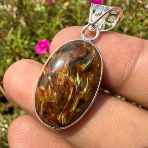 May include: Oval-shaped pendant with a silver bail. The pendant features a polished gemstone with swirling patterns of brown, gold, and black. The gemstone is set in a silver bezel. The background is blurred with pink flowers and green foliage.