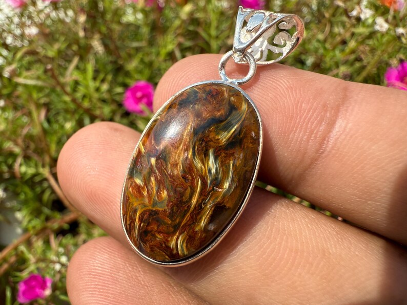 May include: Oval-shaped pendant with a silver bail. The pendant features a polished gemstone with swirling patterns of brown, gold, and black. The gemstone is set in a silver bezel. The background is blurred with pink flowers and green foliage.