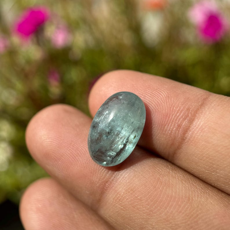 May include: A light blue oval-shaped gemstone with a smooth, polished surface. The gemstone has a translucent appearance with visible internal inclusions.