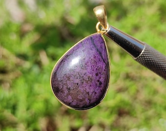 Pode incluir: Um pingente em forma de lágrima com uma pedra preciosa roxa e preta engastada em uma moldura de metal dourado.