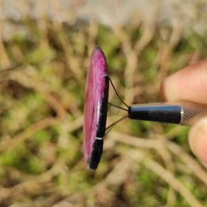 May include: A circular gemstone with a pink interior and a black edge, held by a metal tool. The polished stone displays a contrast of colours and textures. The background is blurred, focusing attention on the gem's details and shape.