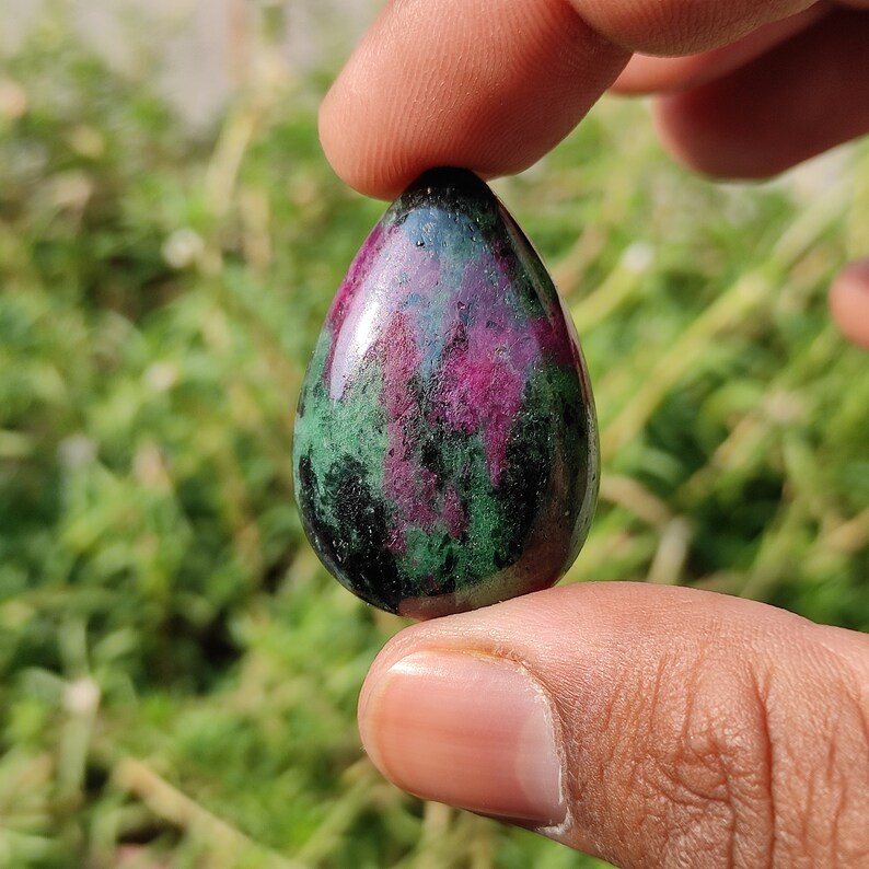 May include: A teardrop-shaped green and pink gemstone pendant. The stone has a smooth, polished surface and is set in a silver tone setting.