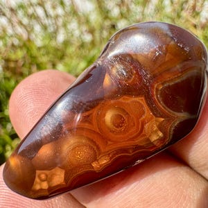 May include: A polished, teardrop-shaped fire agate gemstone featuring deep reddish-brown tones and translucent, layered orbicular patterns that create a glowing, multi-dimensional effect across the smooth, reflective surface of the stone.
