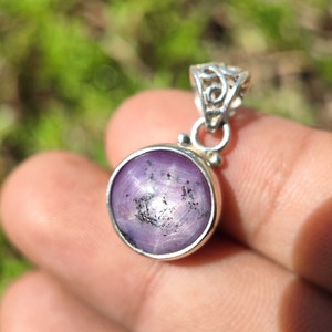 May include: A silver pendant featuring a round, purple star sapphire gemstone with a six-pointed star in the centre. The pendant has a delicate, ornate bail.