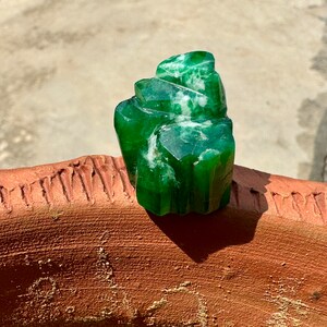 May include: A large, green, faceted crystal with white veins. The crystal is sitting on a brown, textured surface.