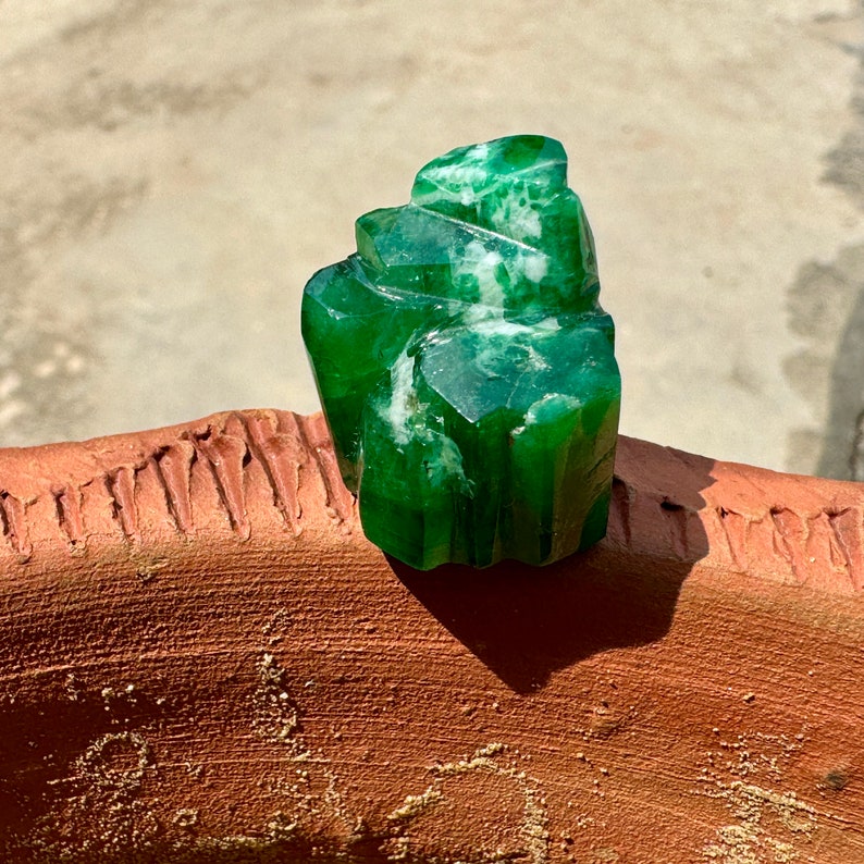 May include: A large, green, faceted crystal with white veins. The crystal is sitting on a brown, textured surface.