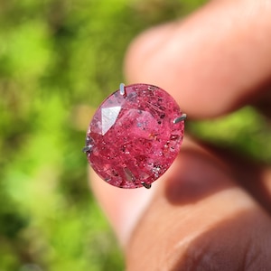 Puede incluir: Una piedra preciosa ovalada de color rosa con un corte facetado. La piedra preciosa está sujeta en su lugar por un engaste de garras de metal.