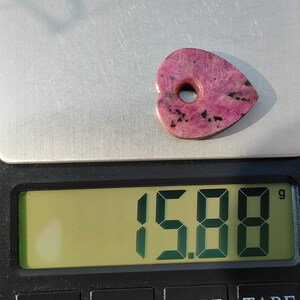 Op de afbeelding: Een roze hartvormige steen met een gat in het midden, met een gewicht van 15,88 gram, op een digitale weegschaal.