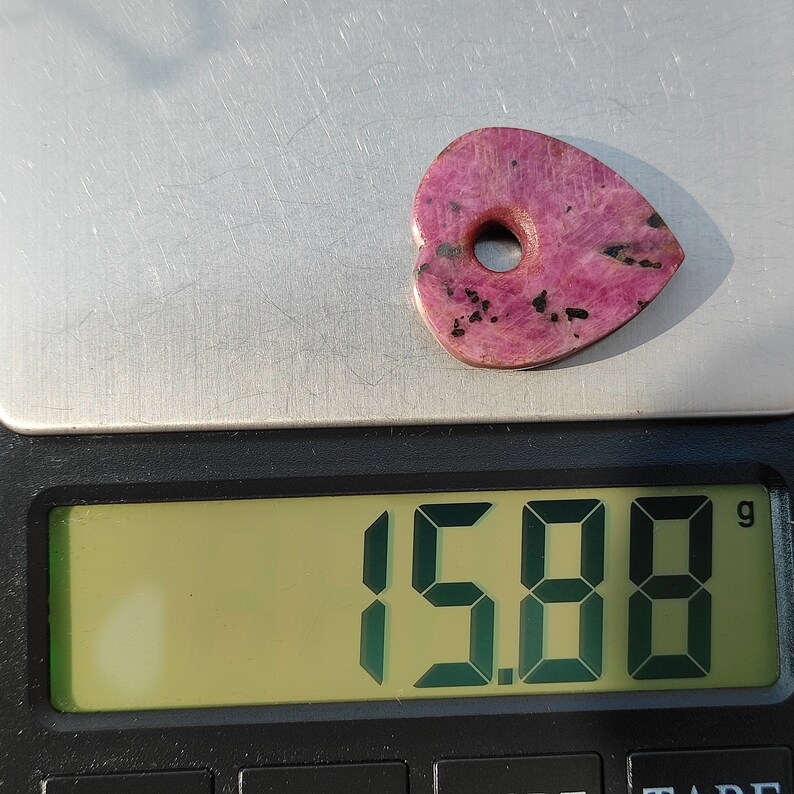 Op de afbeelding: Een roze hartvormige steen met een gat in het midden, met een gewicht van 15,88 gram, op een digitale weegschaal.