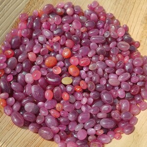 May include: A collection of polished, oval-shaped gemstones in shades of pink and red. The gemstones vary in size and are scattered across a light brown surface, creating a textured appearance. The gems have a glossy finish.