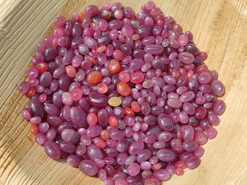 May include: A collection of polished, oval-shaped gemstones in shades of pink and red. The gemstones vary in size and are scattered across a light brown surface, creating a textured appearance. The gems have a glossy finish.