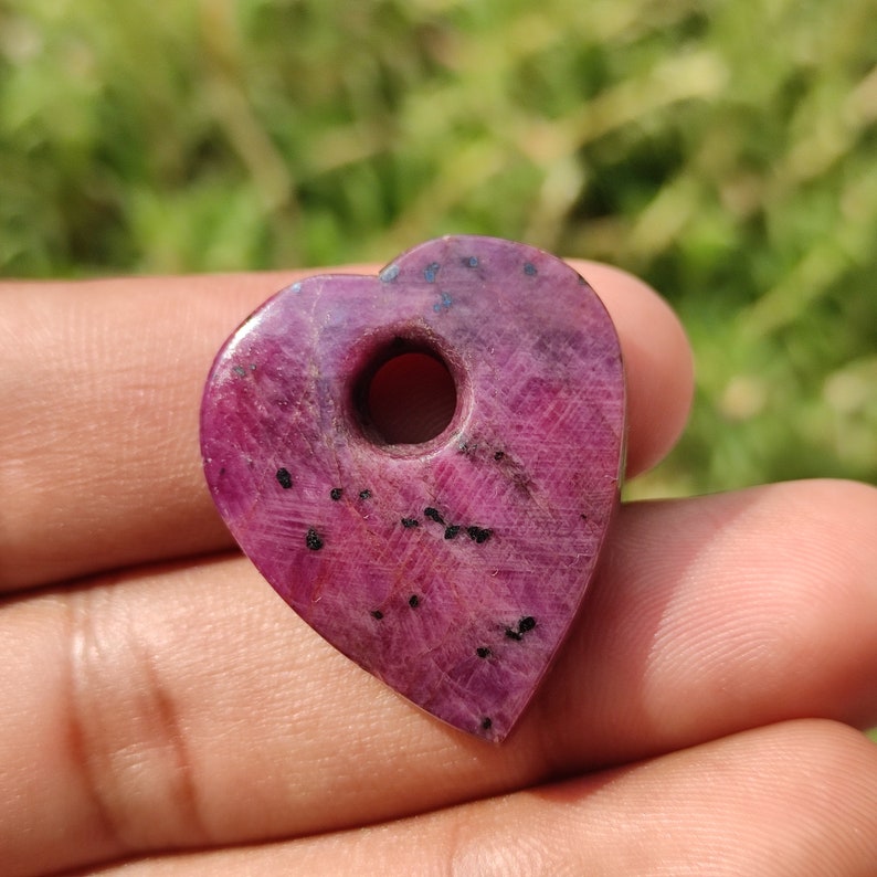 May include: A heart-shaped pendant made of pink ruby with black inclusions. The pendant has a hole drilled through the top for stringing.