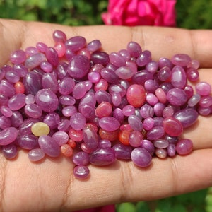 May include: A handful of polished, oval-shaped gemstones in varying shades of pink and purple. The gems are smooth and glossy, with a range of sizes. The gemstones are a mix of light and dark hues, creating a visually appealing display.
