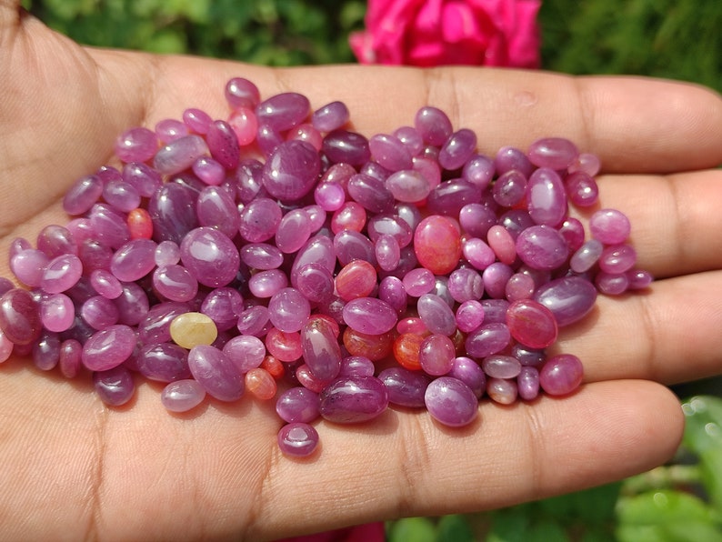 May include: A handful of polished, oval-shaped gemstones in varying shades of pink and purple. The gems are smooth and glossy, with a range of sizes. The gemstones are a mix of light and dark hues, creating a visually appealing display.