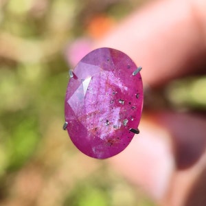 May include: A vibrant pink oval-shaped gemstone with a faceted cut. The gemstone has a slightly cloudy appearance with small black inclusions.