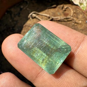Op de afbeelding: Een rechthoekige, doorschijnende edelsteen met een lichtgroene tint. De edelsteen heeft een facetgeslepen slijpsel, met zichtbare interne structuren en subtiele gele en blauwe kleurvariaties. De steen wordt in een hand gehouden, met een vage achtergrond.
