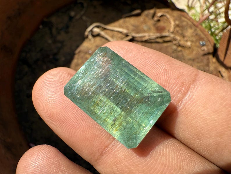 Op de afbeelding: Een rechthoekige, doorschijnende edelsteen met een lichtgroene tint. De edelsteen heeft een facetgeslepen slijpsel, met zichtbare interne structuren en subtiele gele en blauwe kleurvariaties. De steen wordt in een hand gehouden, met een vage achtergrond.