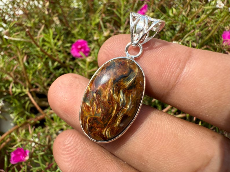 May include: Oval pendant with a polished tiger's eye stone, showcasing swirling patterns of brown, gold, and black. The stone is set in a silver-colored metal frame with a decorative bail. The pendant is held against a background of green foliage and pink flowers.