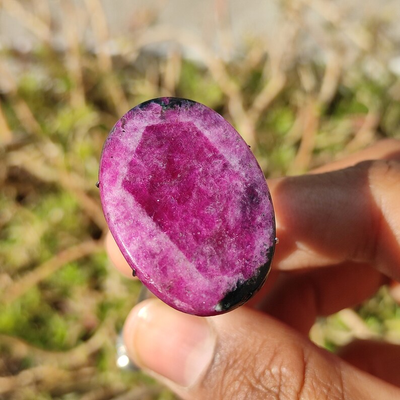 May include: A large, oval gemstone in a vibrant magenta colour. The stone has a faceted, geometric design in the centre, framed by a darker, black border. The gemstone is held in a hand, with a blurred green and brown background.