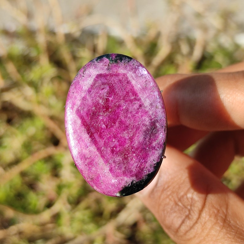 May include: Oval-shaped, polished gemstone with a vibrant magenta hue and a central geometric pattern. The gem has a black border and is held by a person's hand. The background is blurred, suggesting an outdoor setting.