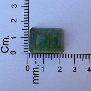 Op de afbeelding: Een rechthoekige, gefacetteerde edelsteen met een lichtgroene tint. De edelsteen is naast een liniaal geplaatst, met metingen in centimeters en millimeters. De edelsteen is ongeveer 2 cm lang.
