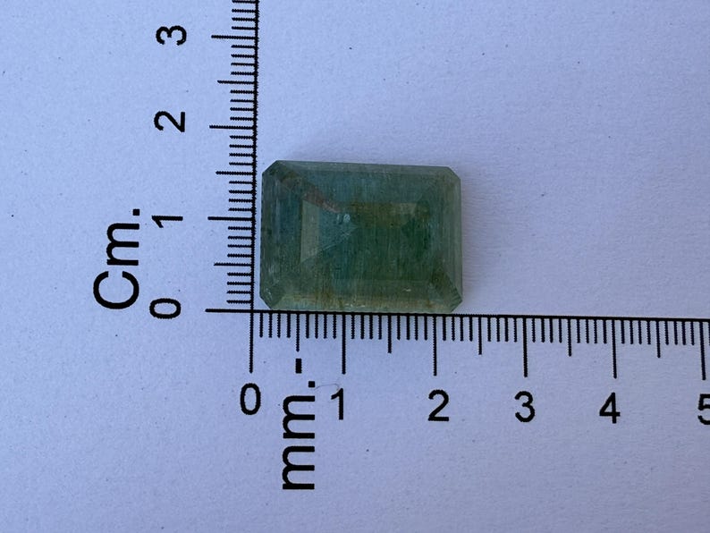 Op de afbeelding: Een rechthoekige, gefacetteerde edelsteen met een lichtgroene tint. De edelsteen is naast een liniaal geplaatst, met metingen in centimeters en millimeters. De edelsteen is ongeveer 2 cm lang.