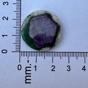 K&ouml;nnte beinhalten: Ein runder, polierter Stein mit einem zentralen lila Muster, umgeben von einem gr&uuml;nen und wei&szlig;en Rand. Der Stein hat einen Durchmesser von etwa 2 cm, wie auf dem Lineal angegeben. Das Design erinnert an einen Edelstein oder ein dekoratives Element.