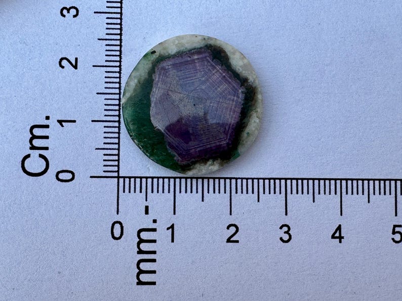 K&ouml;nnte beinhalten: Ein runder, polierter Stein mit einem zentralen lila Muster, umgeben von einem gr&uuml;nen und wei&szlig;en Rand. Der Stein hat einen Durchmesser von etwa 2 cm, wie auf dem Lineal angegeben. Das Design erinnert an einen Edelstein oder ein dekoratives Element.