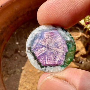 K&ouml;nnte beinhalten: Ein runder Edelstein mit einem zentralen lila, sternartigen Muster. Der Edelstein ist von einem gr&uuml;nen und wei&szlig;en Rand umgeben. Der Stein wird zwischen Fingern gehalten, was seine polierte Oberfl&auml;che und sein aufwendiges Design zeigt. Ein einzigartiges Schmuckst&uuml;ck.