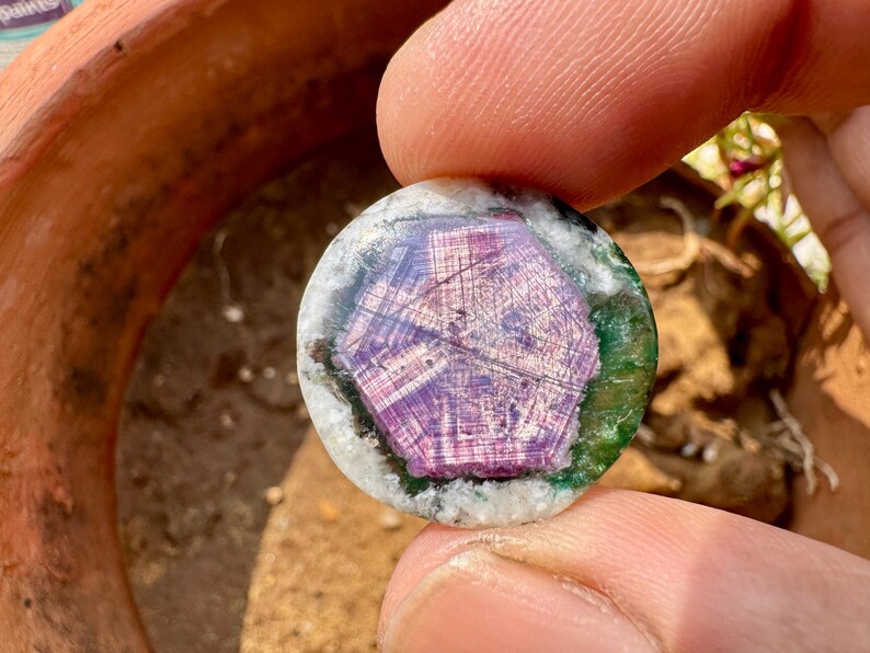 K&ouml;nnte beinhalten: Ein runder Edelstein mit einem zentralen lila, sternartigen Muster. Der Edelstein ist von einem gr&uuml;nen und wei&szlig;en Rand umgeben. Der Stein wird zwischen Fingern gehalten, was seine polierte Oberfl&auml;che und sein aufwendiges Design zeigt. Ein einzigartiges Schmuckst&uuml;ck.