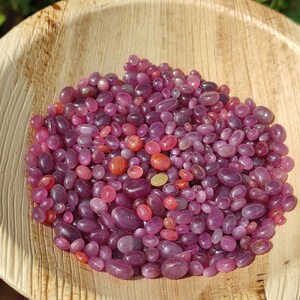 May include: A collection of polished, oval-shaped gemstones in shades of pink and red, filling a natural, light brown, wooden bowl. The gemstones vary in size and color intensity, creating a visually appealing display. The bowl's texture contrasts with the smooth stones.