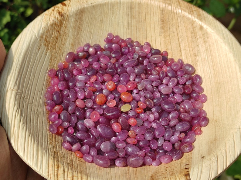 May include: A collection of polished, oval-shaped gemstones in shades of pink and red, filling a natural, light brown, wooden bowl. The gemstones vary in size and color intensity, creating a visually appealing display. The bowl's texture contrasts with the smooth stones.