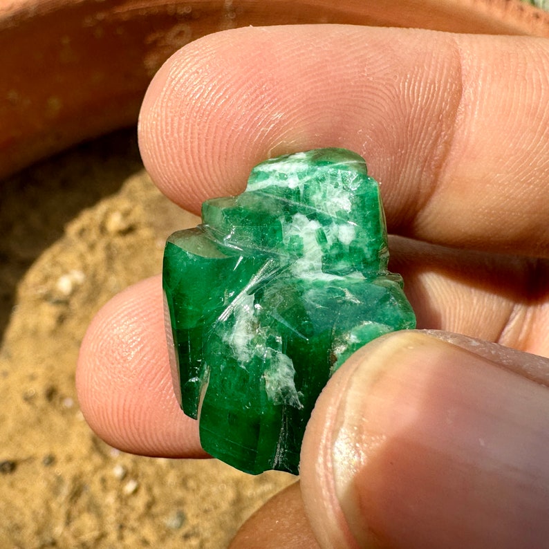 May include: A large, green emerald gemstone with white inclusions. The emerald is carved into a unique shape and has a smooth, polished surface.