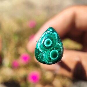 May include: A green malachite cabochon with a swirling pattern of white and green rings. The cabochon is set in a silver bezel and is ready to be used in jewelry making.